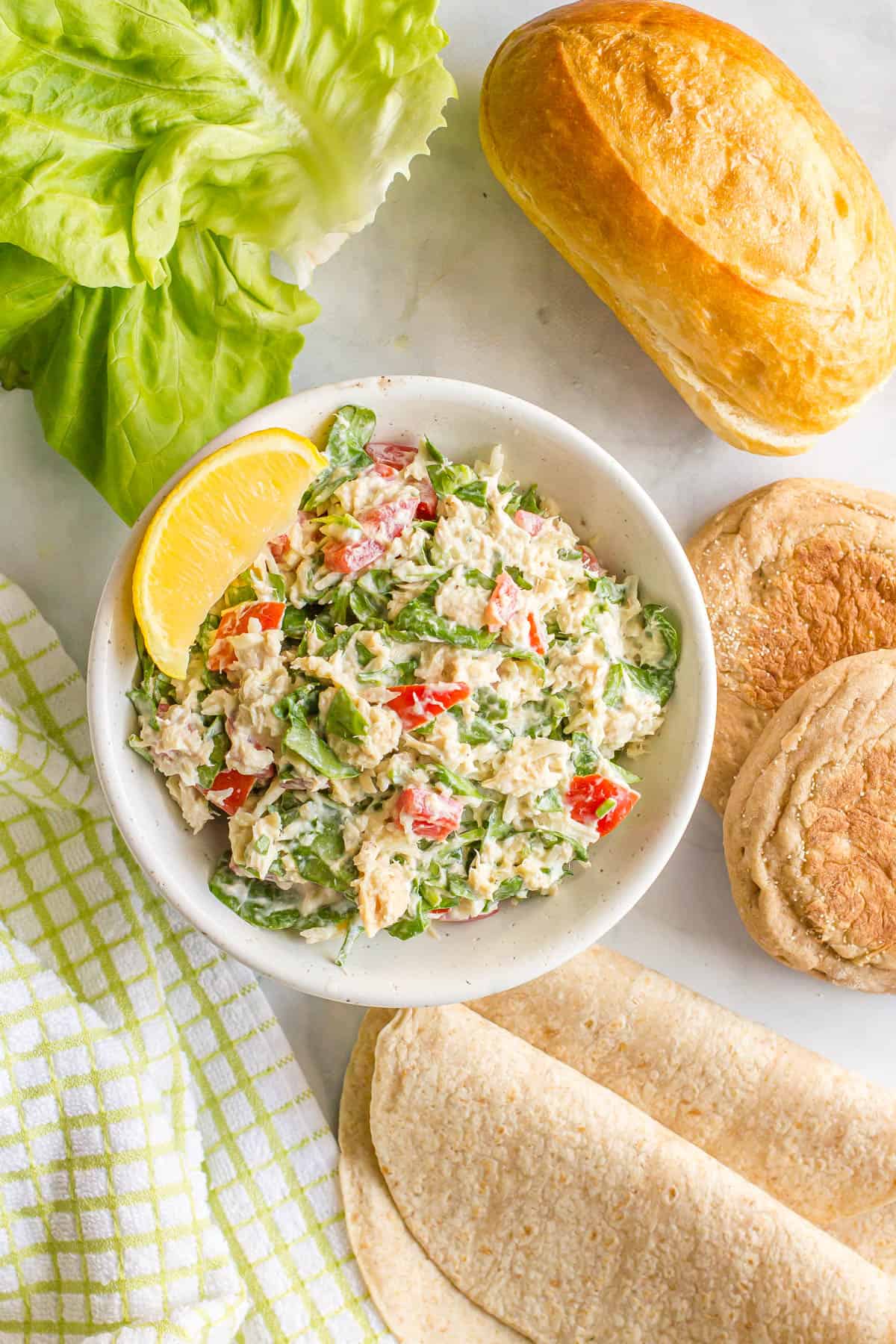 Tuna salad with spinach, tomatoes and mozzarella in a small bowl with bread, wraps, English muffins and Bibb lettuce to the side.