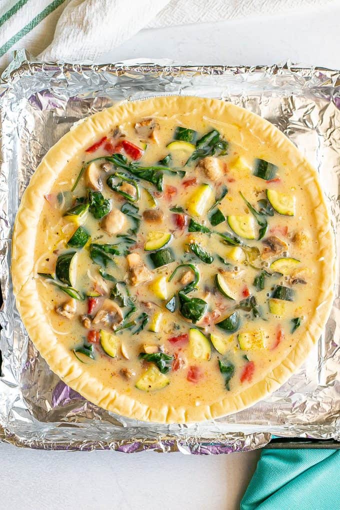 A pie crust set on a foil lined baking tray and filled with an egg and veggie mixture before being cooked.
