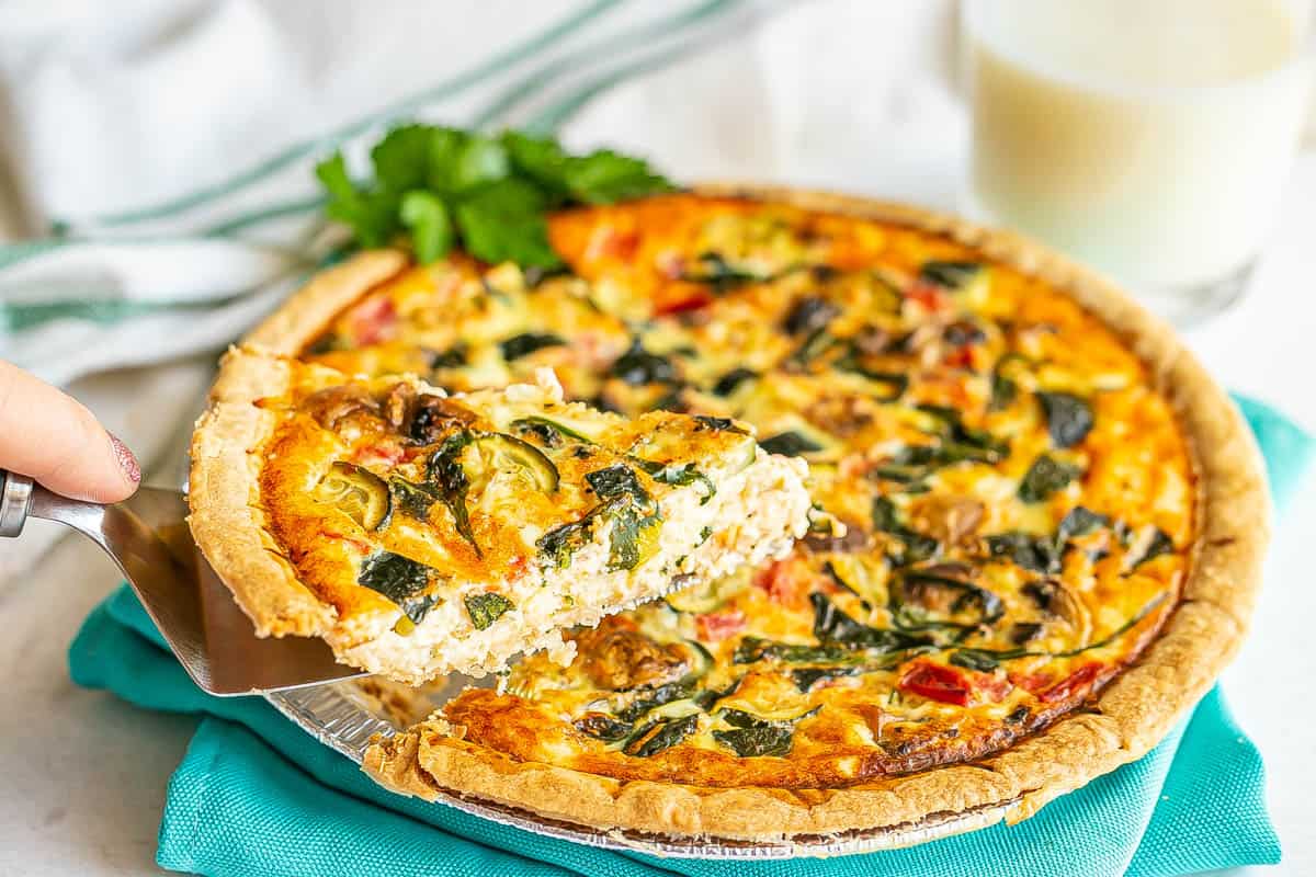 A slice of vegetable quiche with spinach, mushrooms and zucchini being taken out of a pie plate.