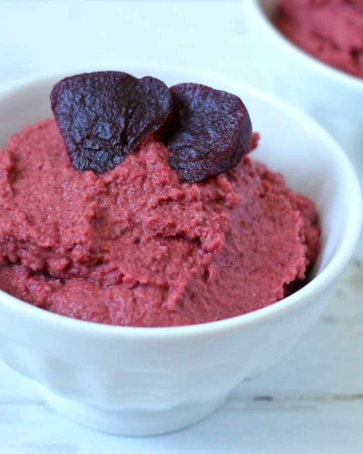 A small white bowl of beet hummus topped with sliced cooked beets.