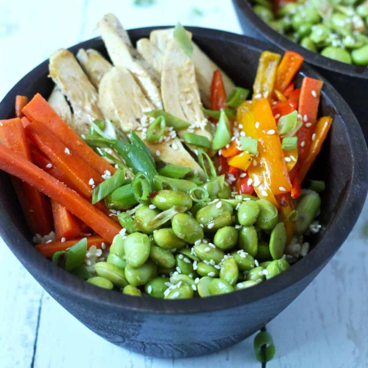 A brown rice bowl with chicken, colorful bell peppers, edamame, carrot sticks and sesame seeds sprinkled on top.