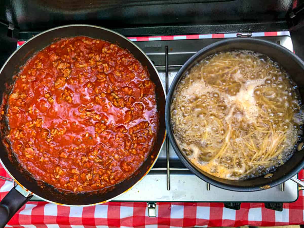 Spaghetti meat and sauce cooking in one pan along side a pot of spaghetti cooking on a camp stove.