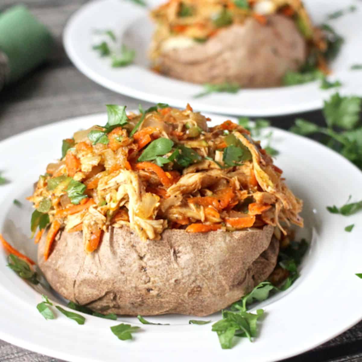 A shredded chicken, onion and carrot hash mixture served over a baked potato in a low white bowl.