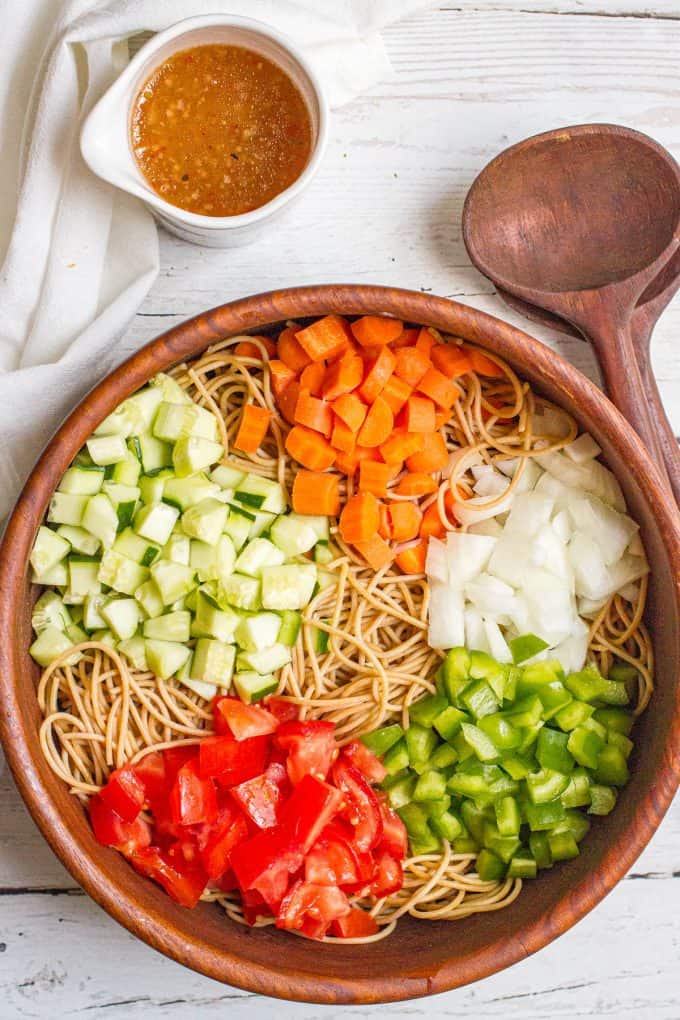 Spaghetti in a large wooden bowl with piles of cucumber, carrots, onion, green pepper and tomatoes before being mixed in.