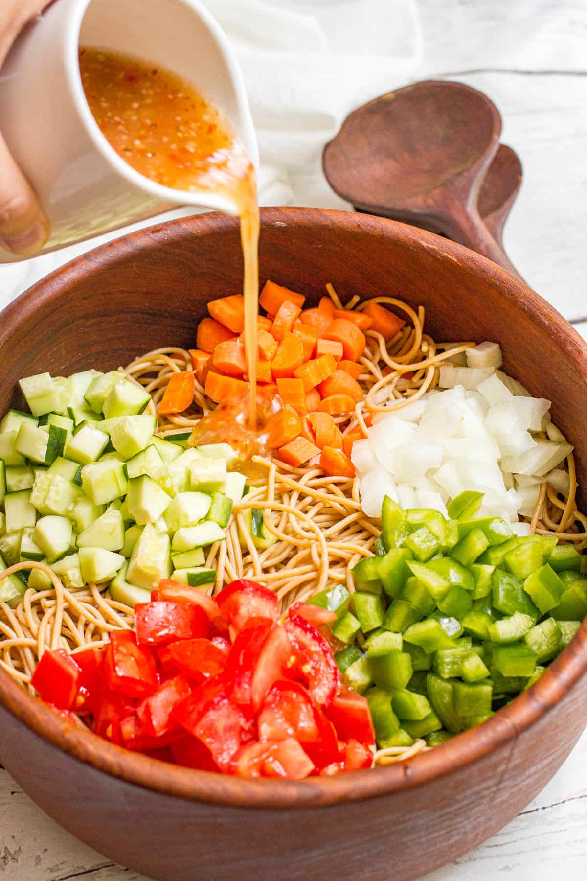 Italian dressing being poured over a spaghetti salad with fresh veggies.