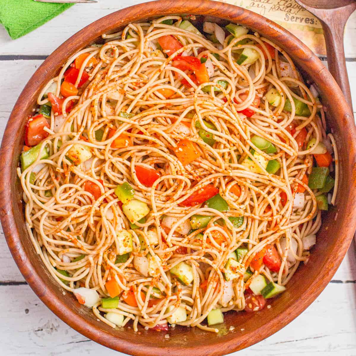 A tossed spaghetti salad with fresh veggies and dressing in a wooden bowl.