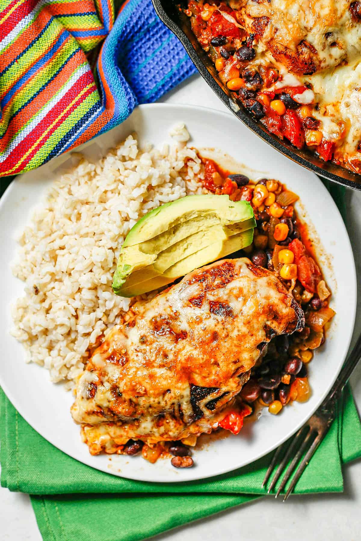 A white dinner plate with cheesy chicken served with beans, corn and tomatoes alongside steamed rice with slices of avocado in the middle.