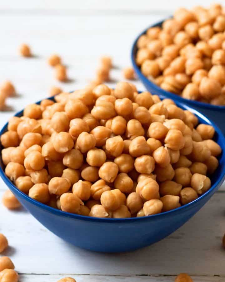 Two blue bowls full of chickpeas cooked in a crock pot with extra chickpeas scattered on the counter.