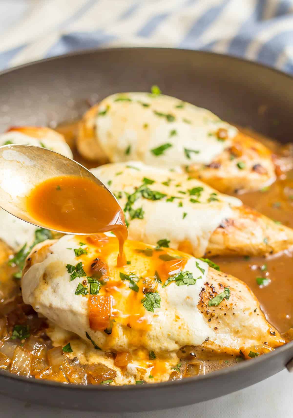 A spoon pouring pan juices over a mozzarella cheese covered chicken breast in a pan after baking.