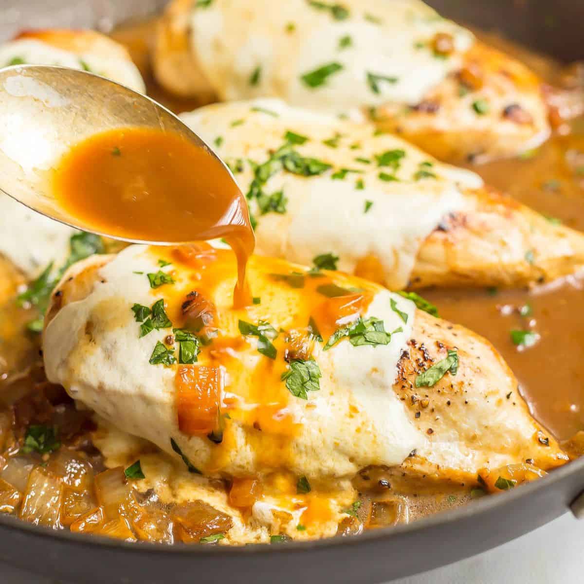 Close up of a spoon pouring pan juices over a mozzarella cheese covered chicken breast in a pan after baking.