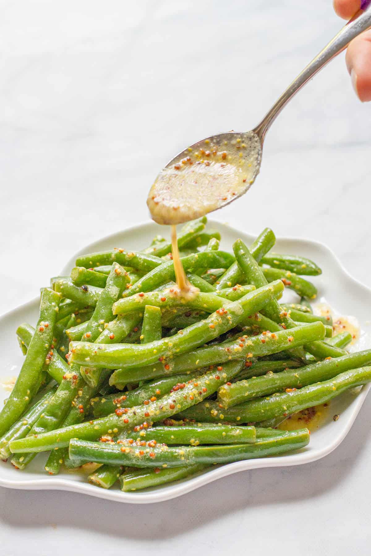 A spoon drizzling some mustard butter sauce over a small serving plate of steamed green beans.