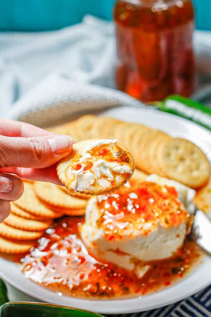 A hand holding a cracker with a spread of cream cheese and pepper jelly on it.