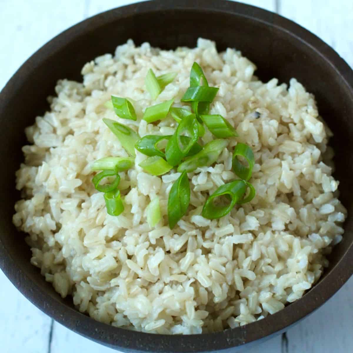 Close up of steamed brown rice in a brown bowl topped with sliced green onions.