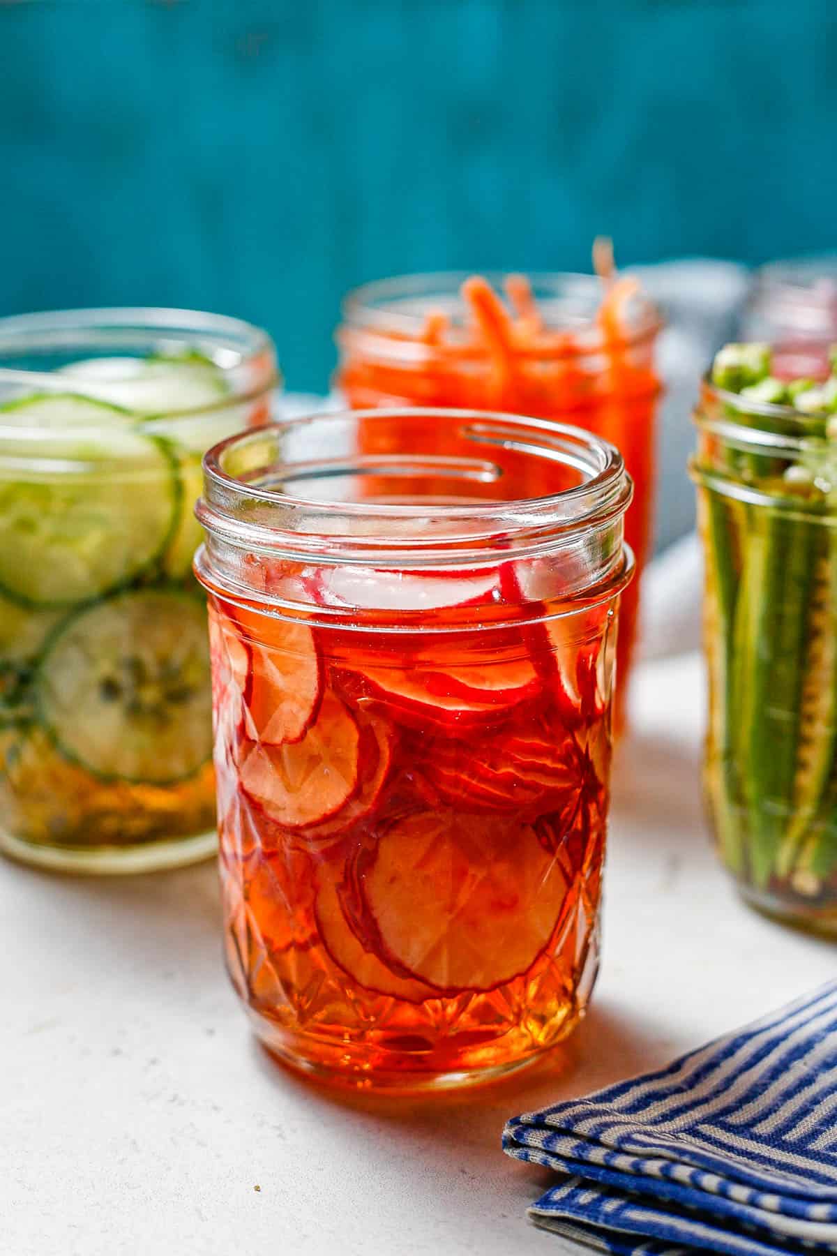Sliced pickled radishes in a small glass jar with other pickled veggies in the background.