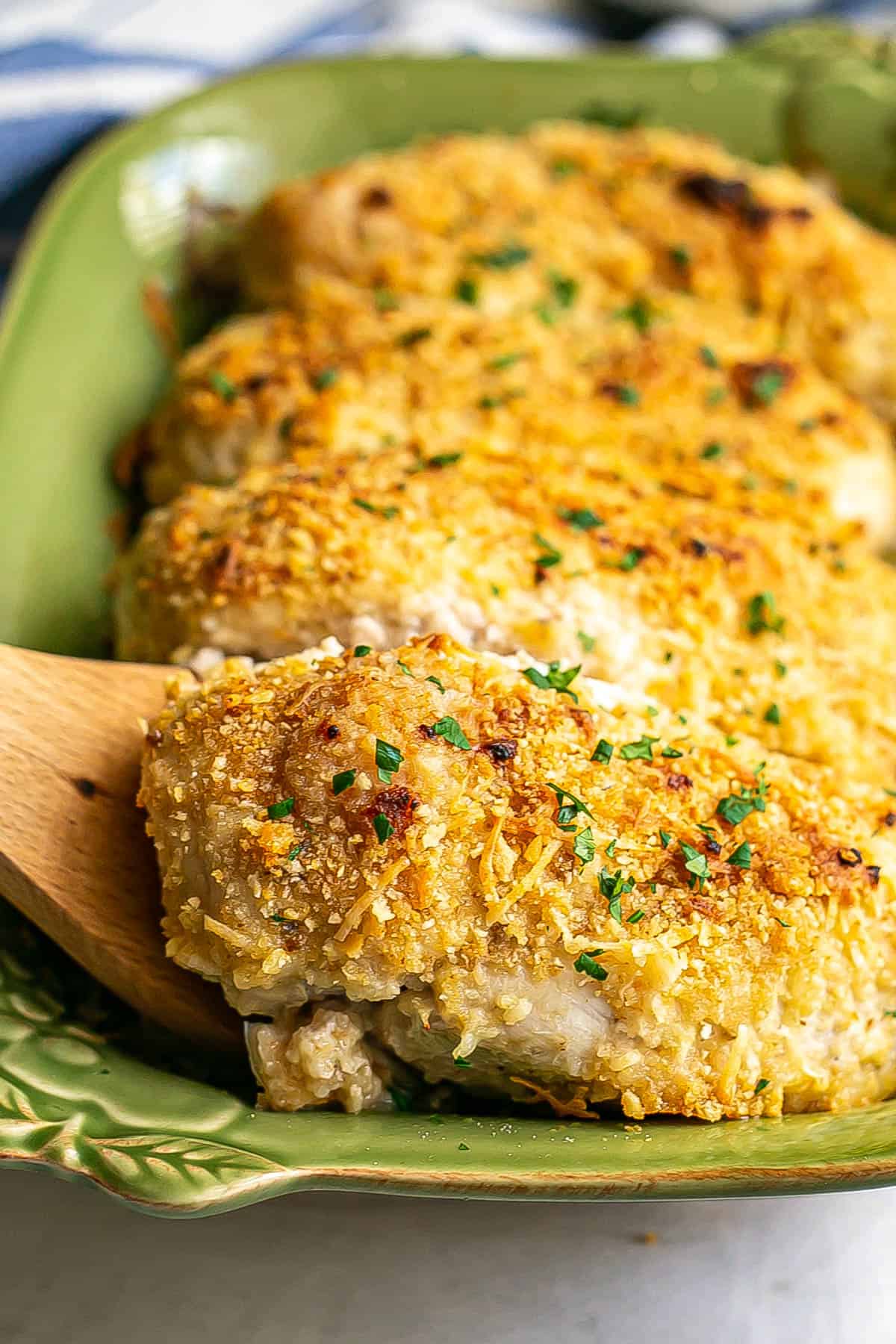 A wooden spatula lifting out a cheesy baked chicken breast from a green casserole dish.