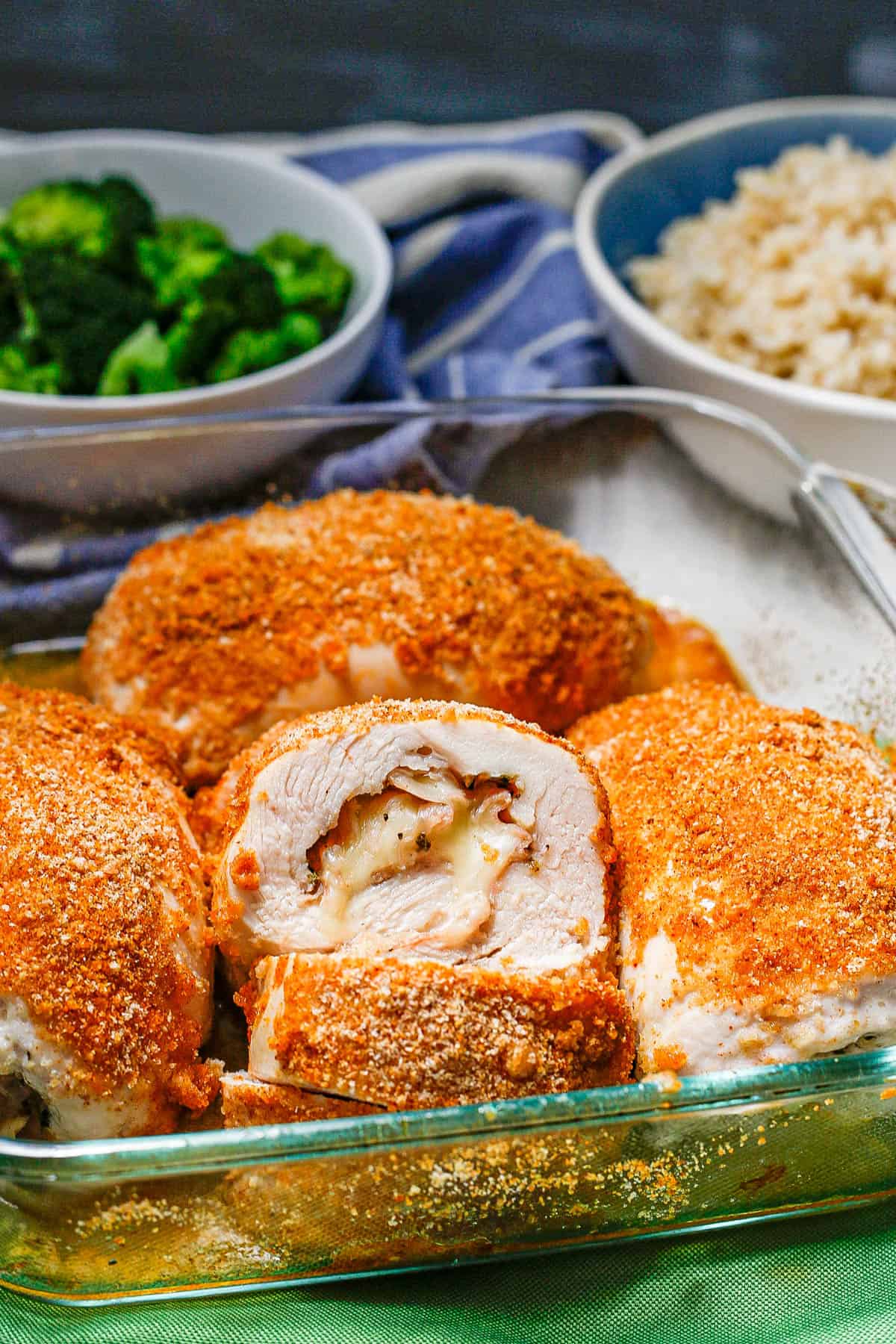 A glass casserole dish of baked chicken cordon bleu with bowls of steamed broccoli and steamed brown rice in the background.