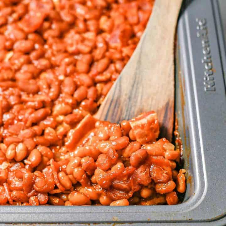 Close up of a scoop of baked beans being taken from a pan with a wooden spatula.
