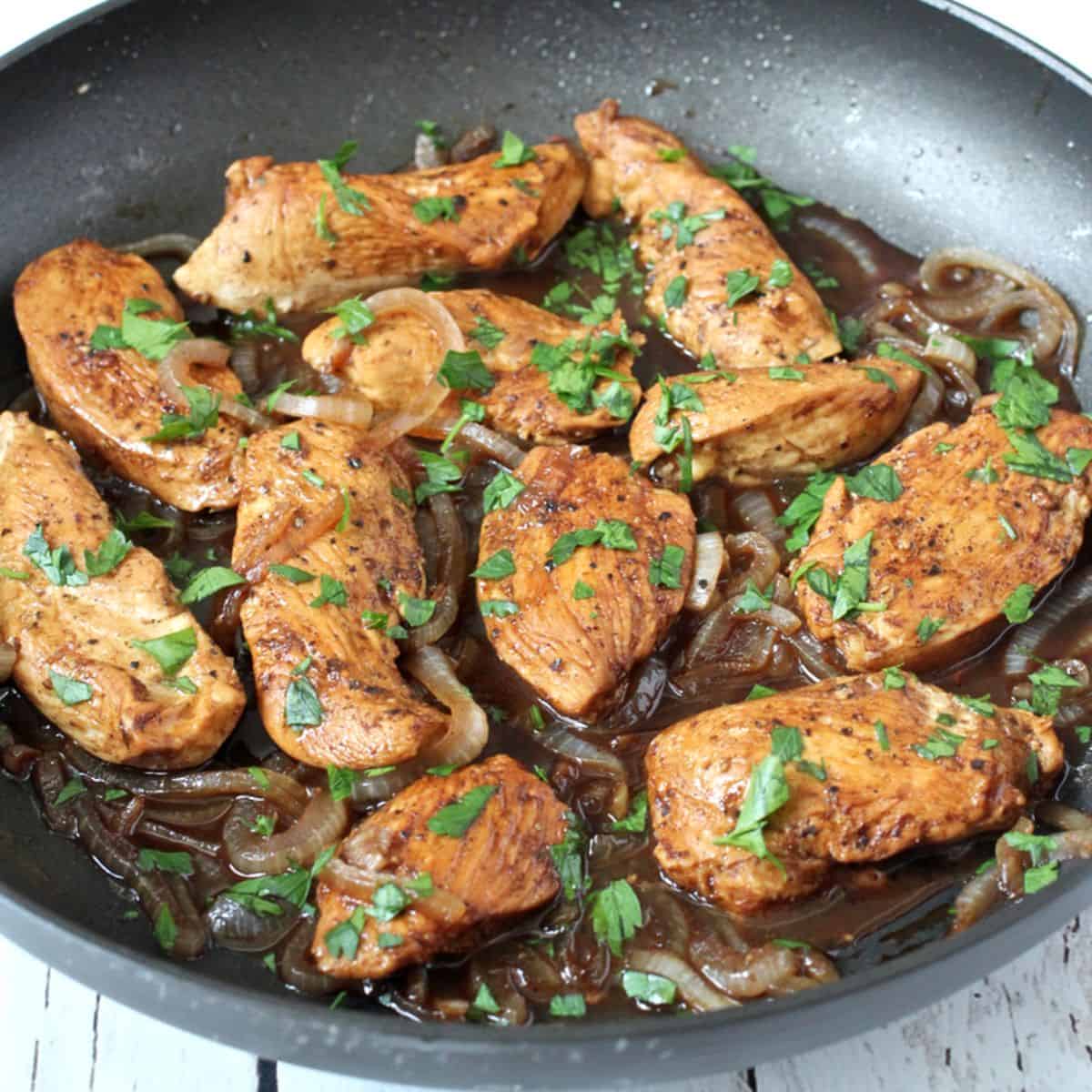 Close up of cooked strips of chicken breast with onions and a balsamic vinegar sauce in a skillet, with fresh parsley sprinkled on top.