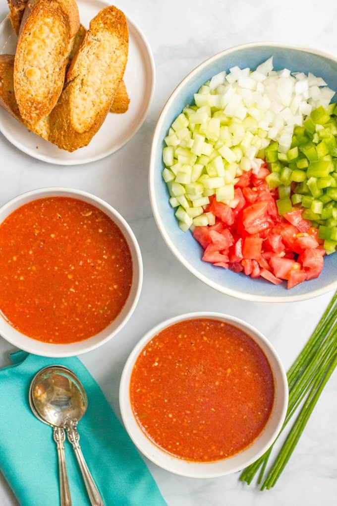 Two bowls of gazpacho soup with a bowl full of chopped veggies that will be stirred in.