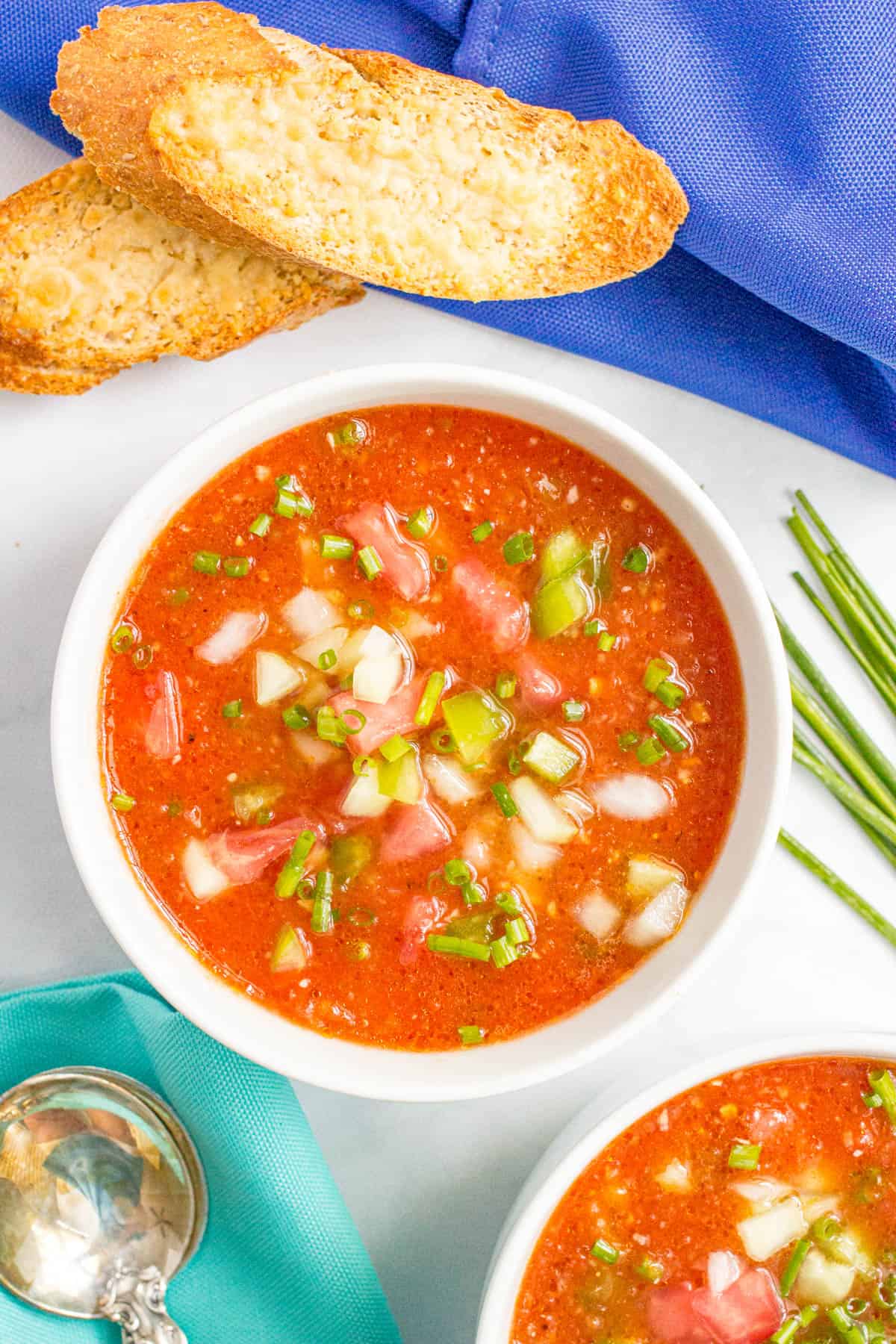 A bowl of gazpacho soup with chives to the side and a couple of toasted baguette pieces nearby.