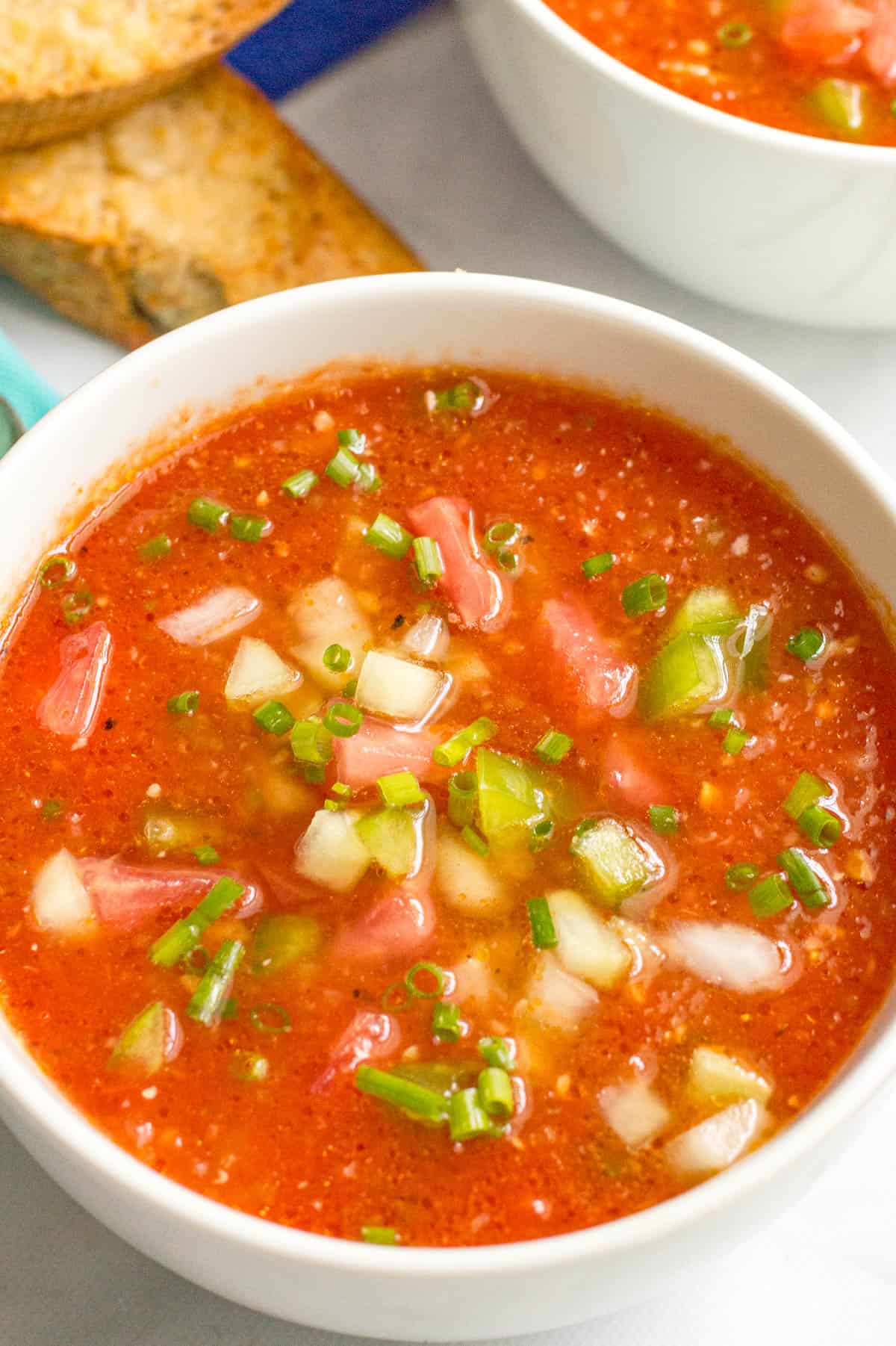 Close up of a white bowl filled with tomato gazpacho soup with fresh veggies and snipped chives on top.