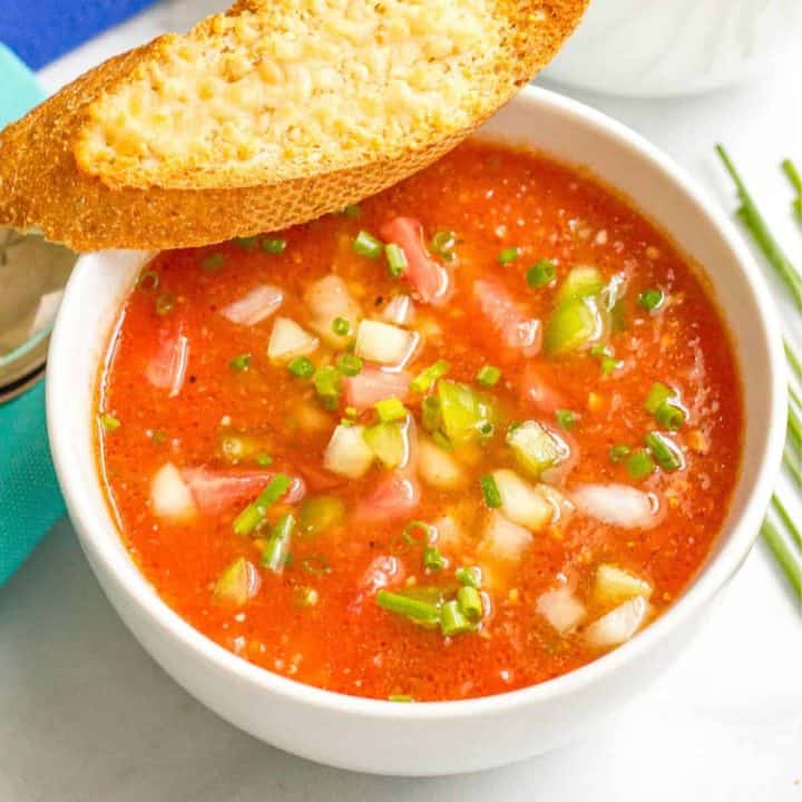 Close up of a bowl of gazpacho with a slice of cheesy baguette resting on the side of the bowl.
