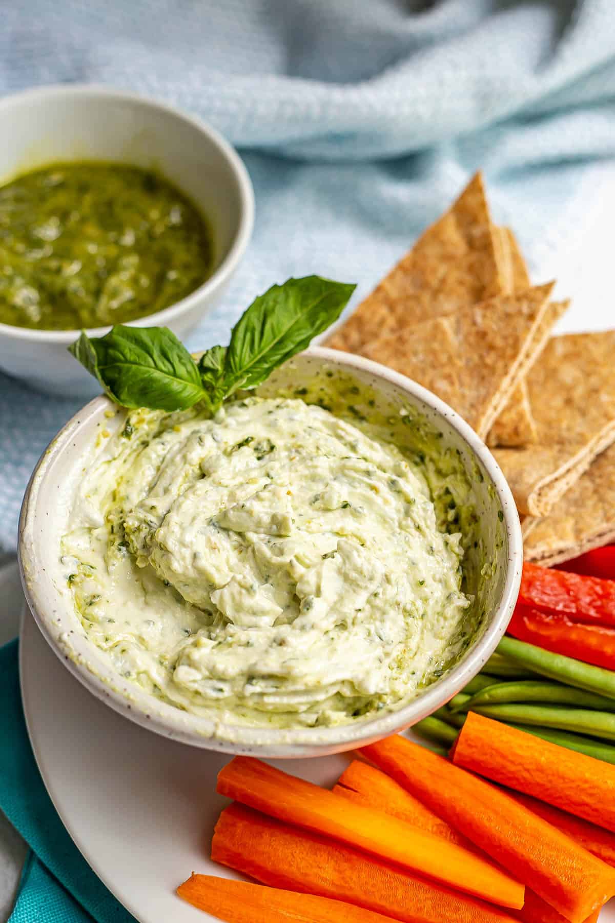 A pesto Greek yogurt dip served in a bowl with a garnish of basil and fresh veggies and pita triangles for dipping.