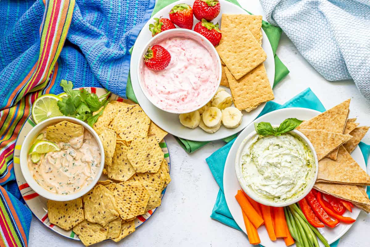 A trio of Greek yogurt dips in bowls on plates with an assortment of foods for dipping.