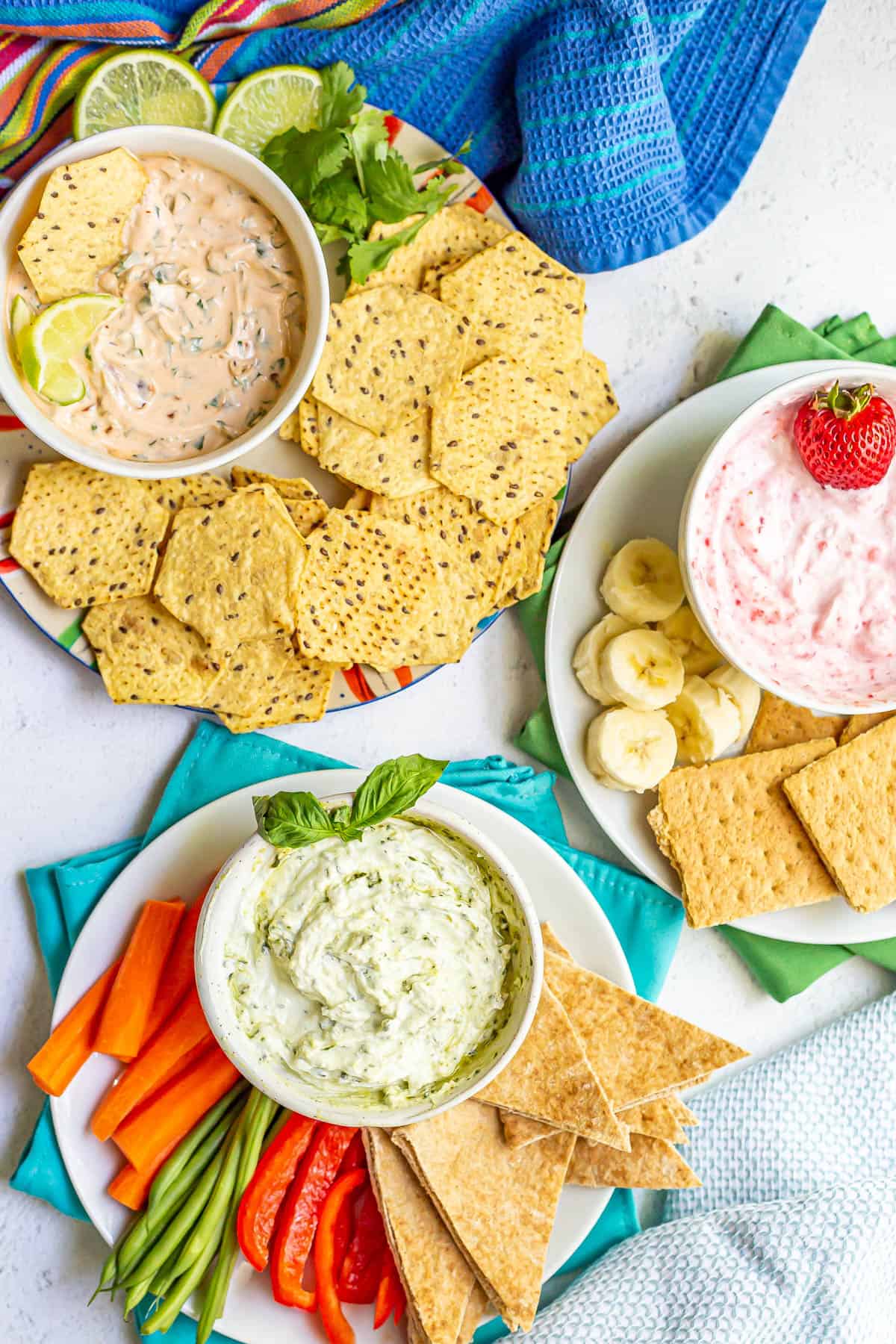 Three different Greek yogurt dips in bowls on plates with an assortment of foods for dipping.