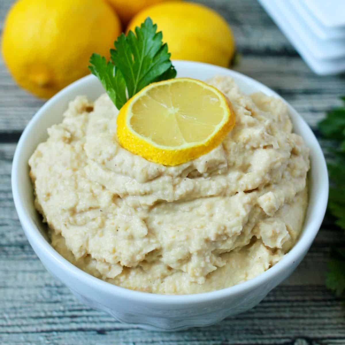 A small white bowl with homemade hummus topped with a small lemon slice and a sprig of parsley.