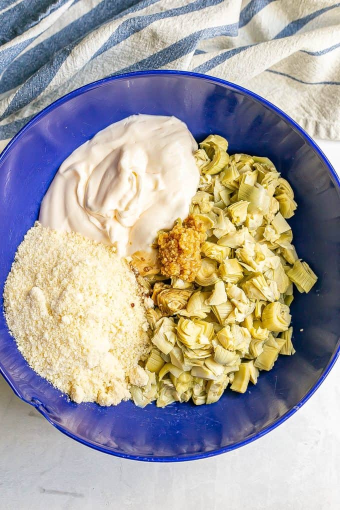 A blue mixing bowl with artichokes, Parmesan cheese and a creamy mixture before being mixed together.