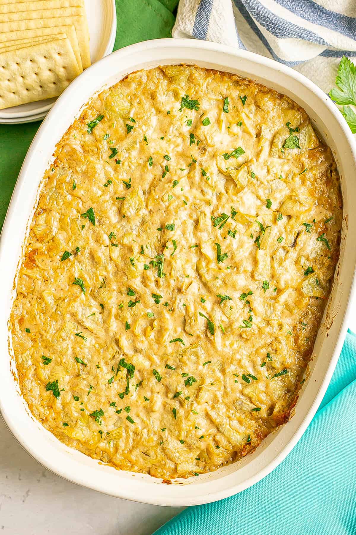 A hot and bubbly baked artichoke dip with parsley on top and crackers to the side.