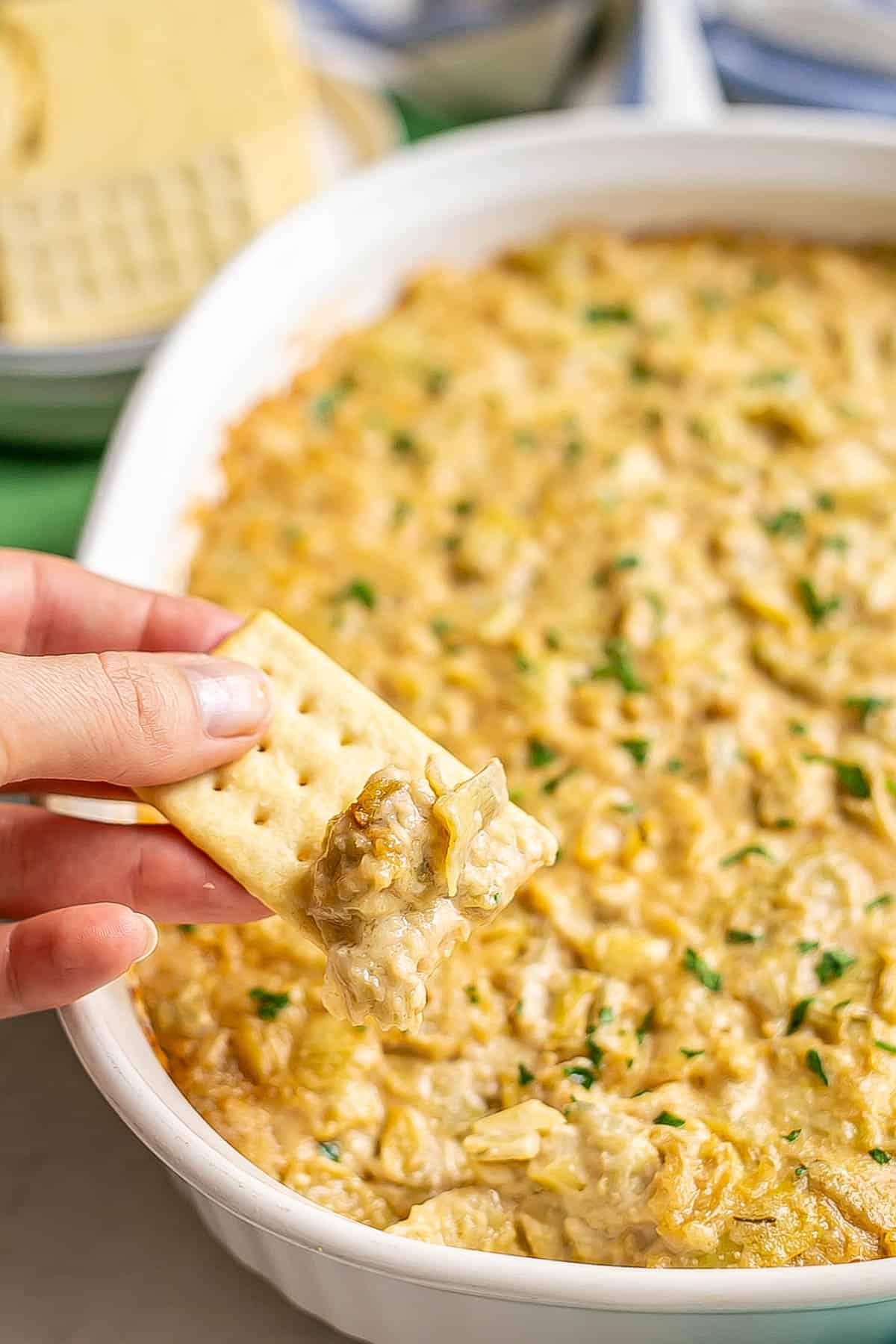 A hand holding up a cracker with a creamy artichoke dip.