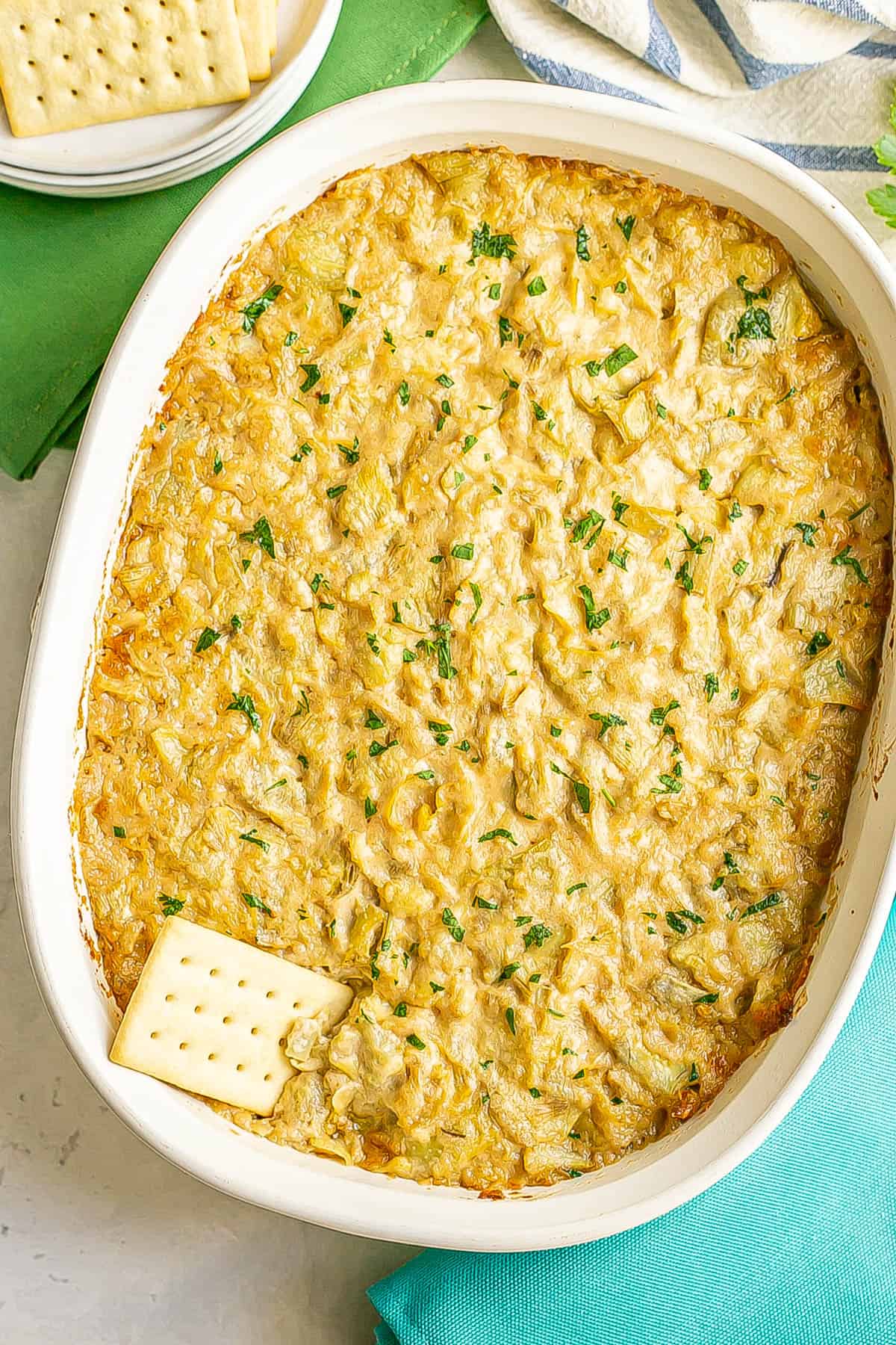 A cracker resting in a white oval casserole dish filled with a creamy artichoke mixture topped with chopped fresh parsley.