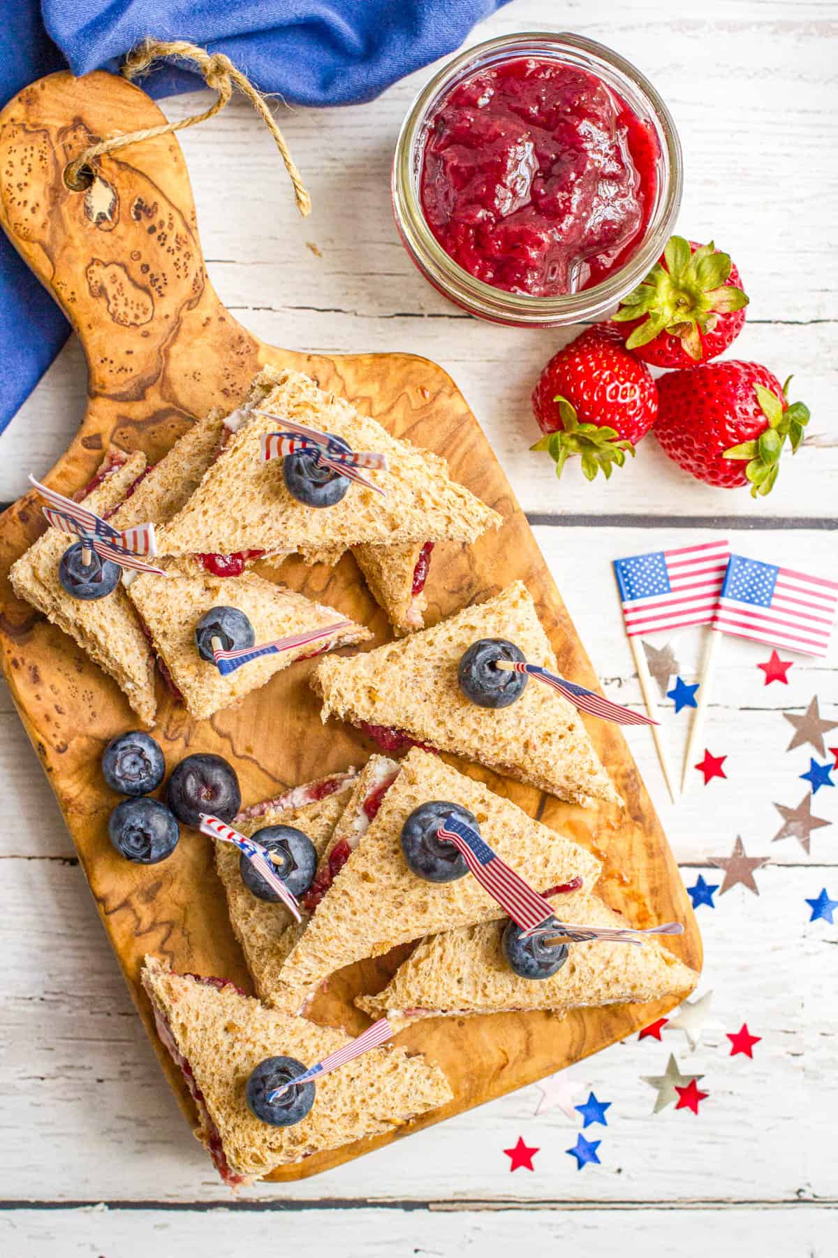 Mini triangle sandwiches stacked on a cutting board with cream cheese, strawberry jam and a blueberry on top held in place with mini American flags.