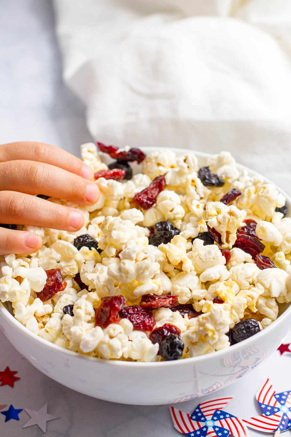 A small hand reaching into a white bowl of popcorn mixed with dried blueberries and dried cranberries.