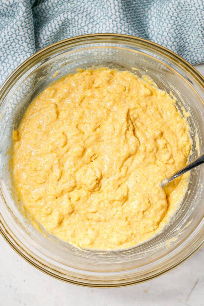 Homemade cornbread batter in a large glass bowl with a spoon resting in it.