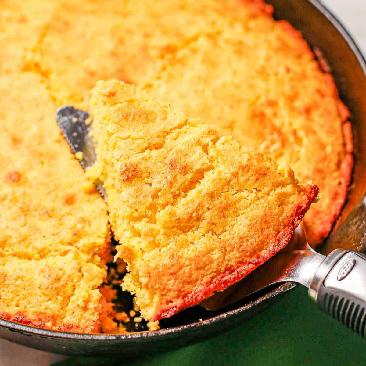 Close up of a pie server lifting up a slice of cornbread from a cast iron skillet.