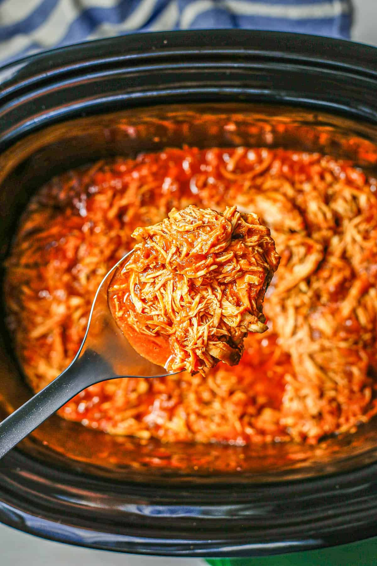 A scoop of slow cooker BBQ chicken being lifted out of the crock pot insert.