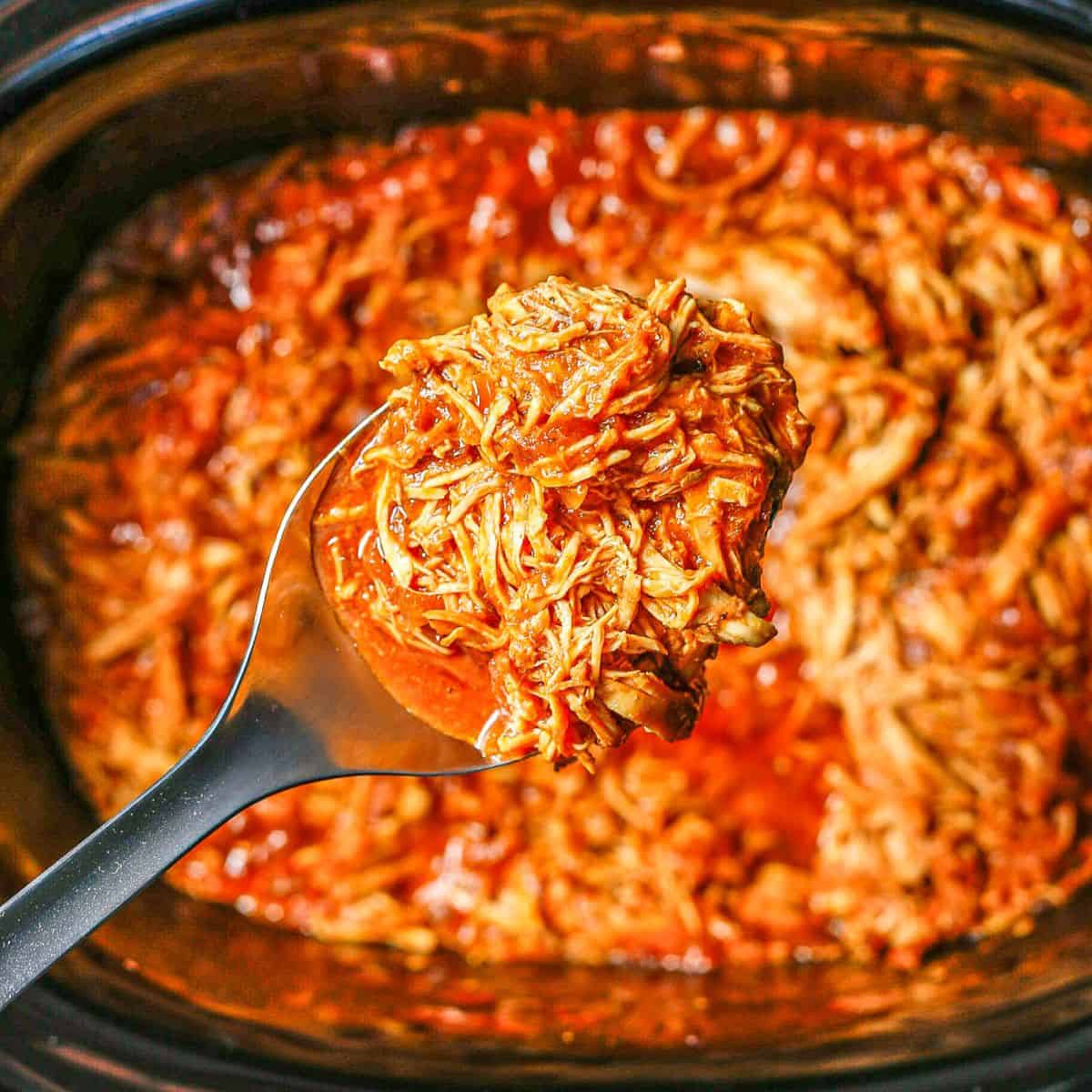 Close up of a scoop of slow cooker BBQ chicken being lifted out of the crock pot insert.