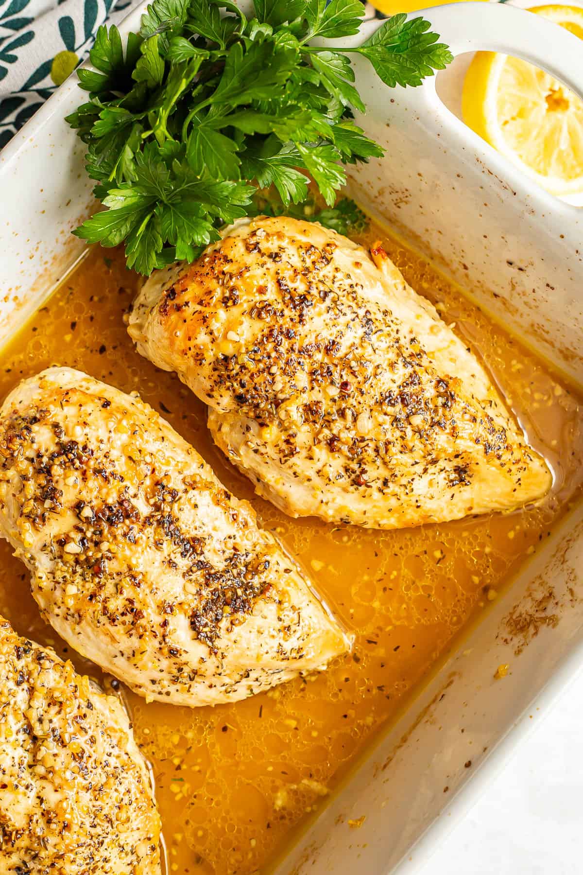 Three seasoned and seared chicken breasts in a white casserole dish with a lemony sauce and sprigs of parsley tucked to the side.