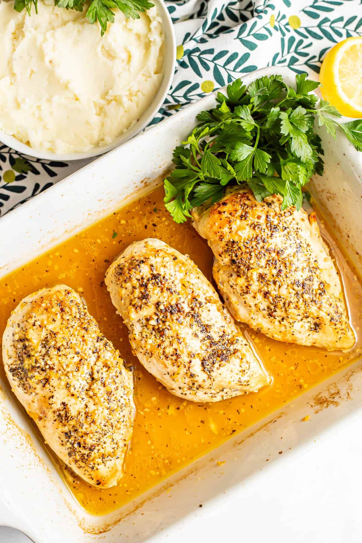 Baked lemon chicken breasts in a white casserole dish with parsley to the side and a bowl of mashed potatoes nearby.