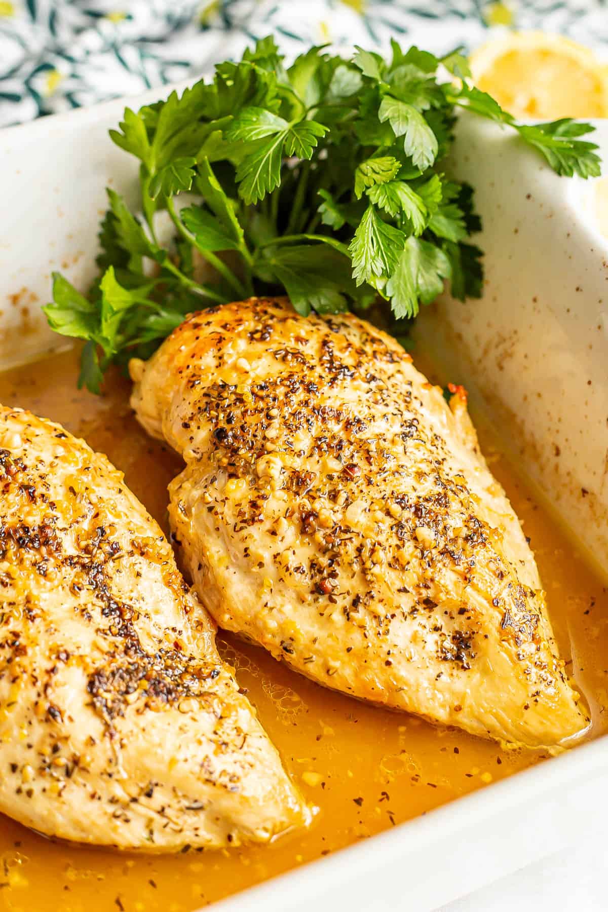 Close up of baked lemon chicken in a white rectangular casserole dish with parsley to the side for garnish.