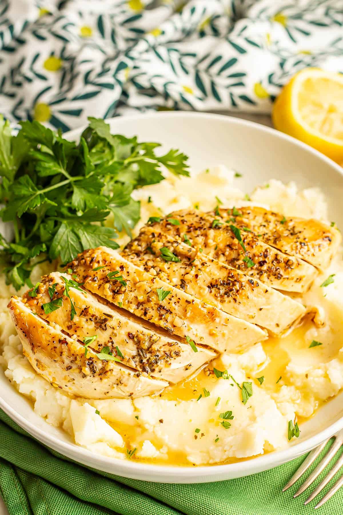 Close up of a sliced chicken breast served on top of mashed potatoes with parsley for a garnish and cut lemons to the side.