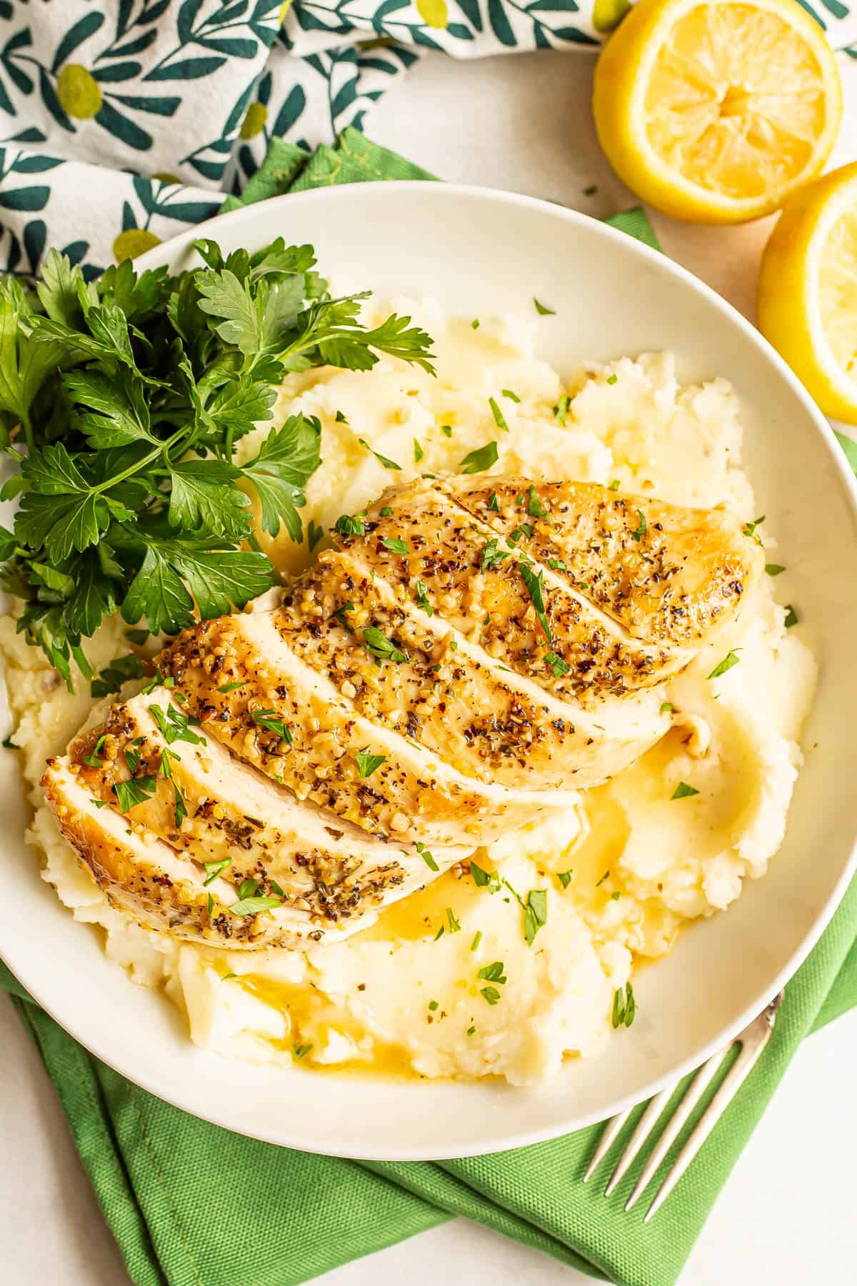 Sliced chicken breast served on top of mashed potatoes with parsley for a garnish and cut lemons to the side.