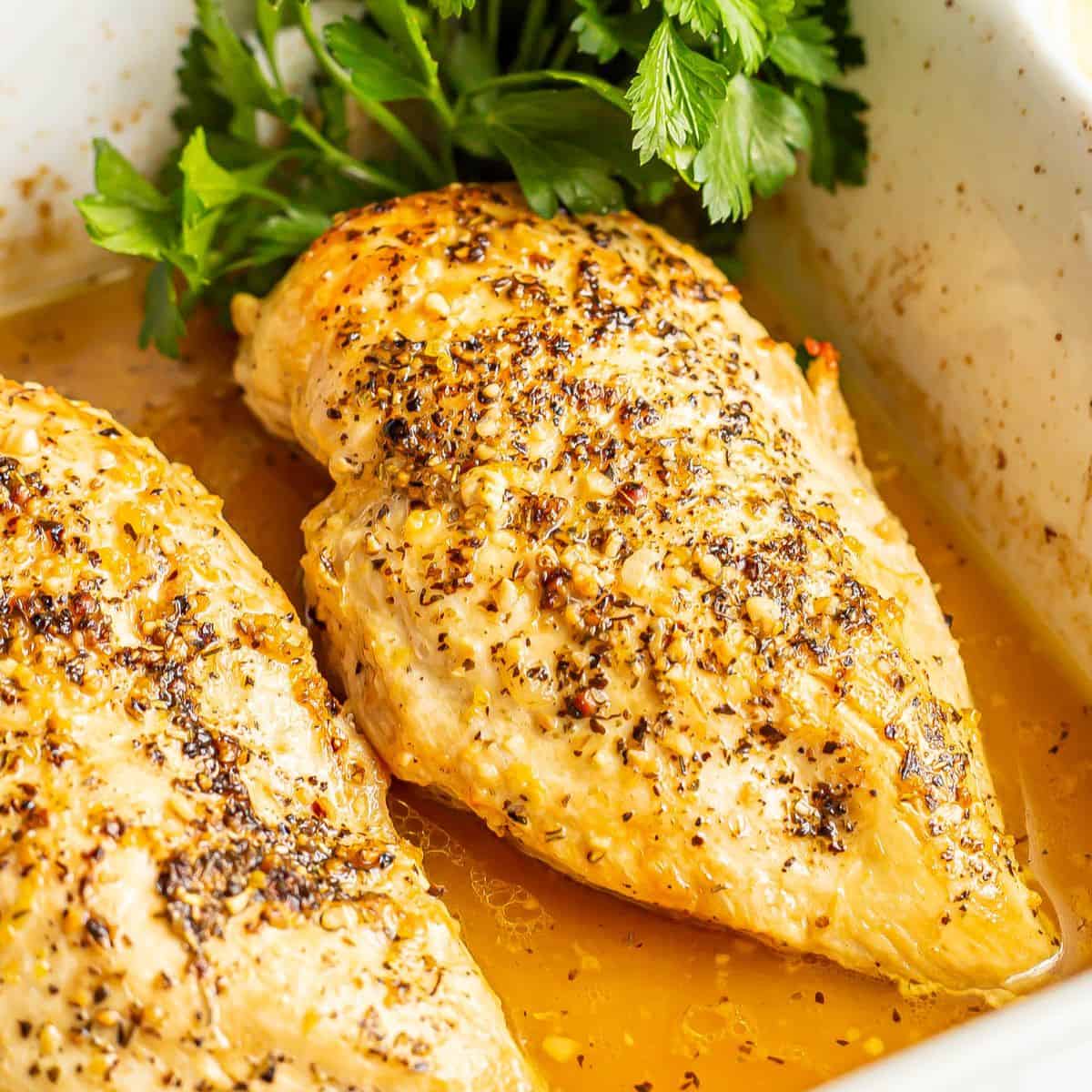 Close up of a baked chicken breast in a white rectangular casserole dish with parsley to the side for garnish.