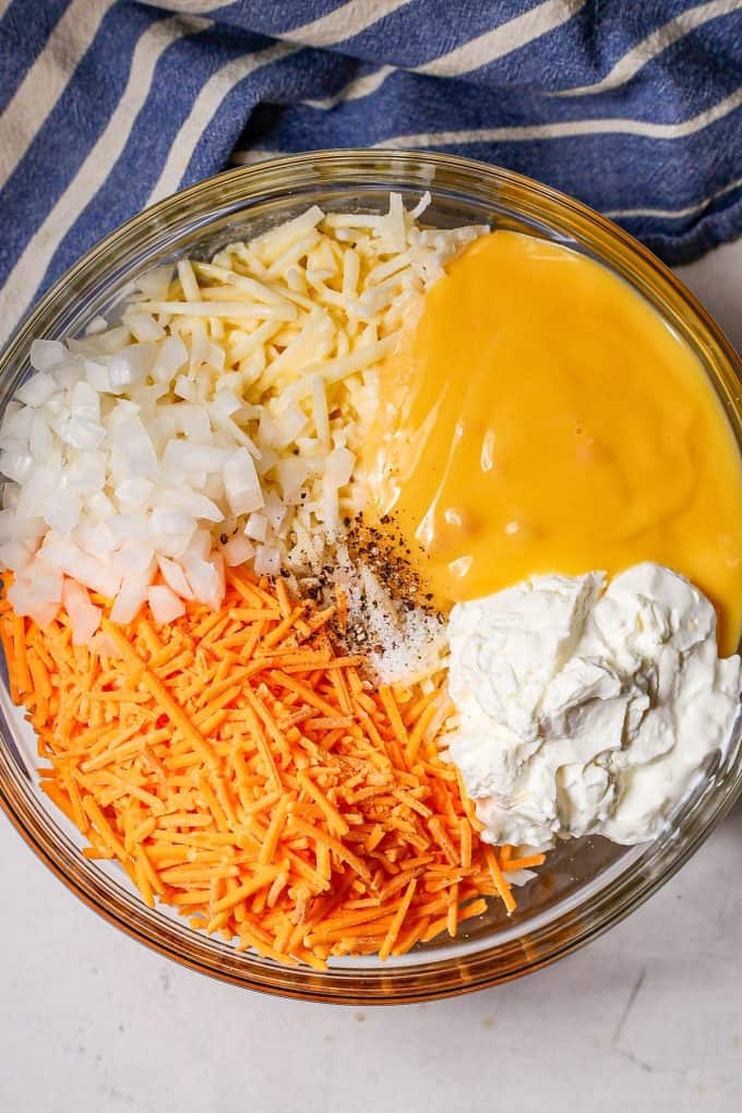 A large glass bowl with hash browns topped by piles of onion, cheese, cream of chicken soup and Greek yogurt.