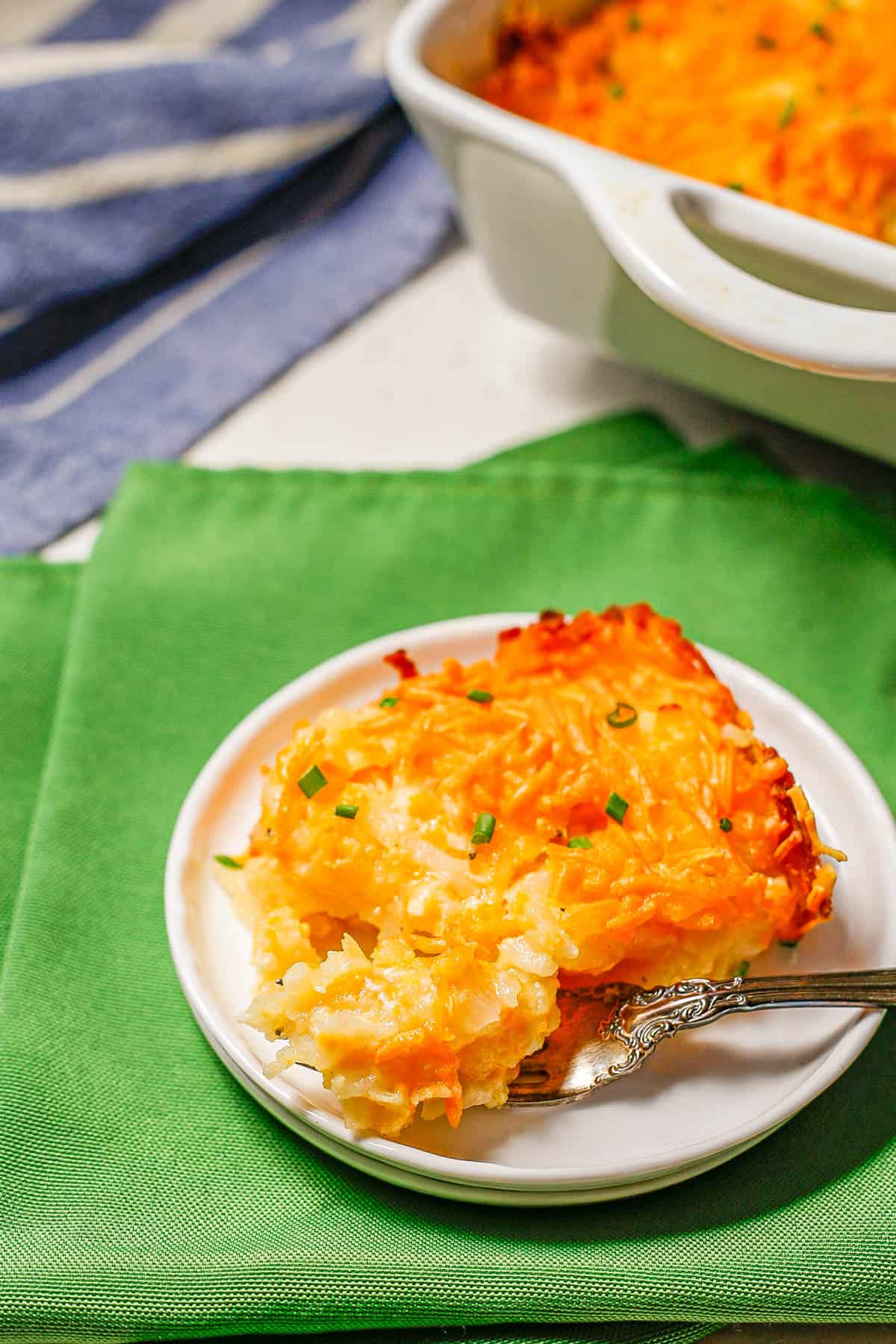 A fork with a bite of cheesy hash brown casserole served on a small white plate set on green napkins.