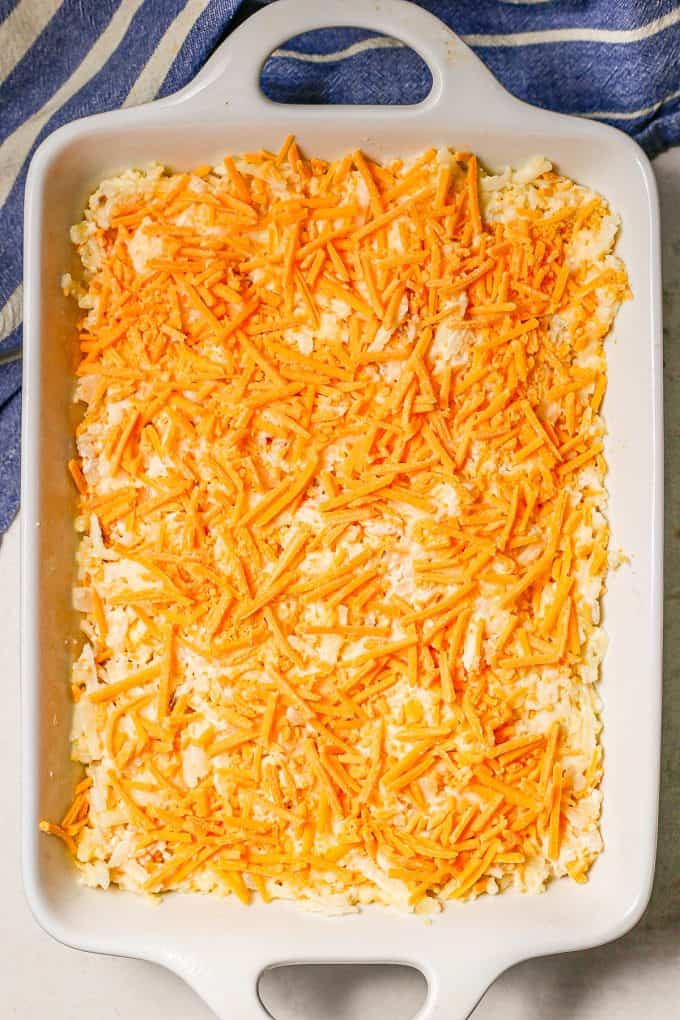 A hash brown casserole topped with shredded cheddar cheese in a large white rectangular casserole dish before being cooked.