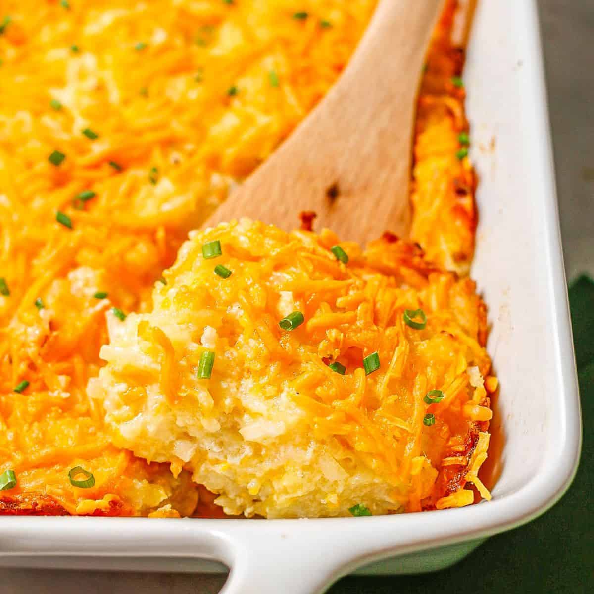 Close up of a wooden spatula lifting out a square of cheesy hash brown casserole from a white rectangular dish.